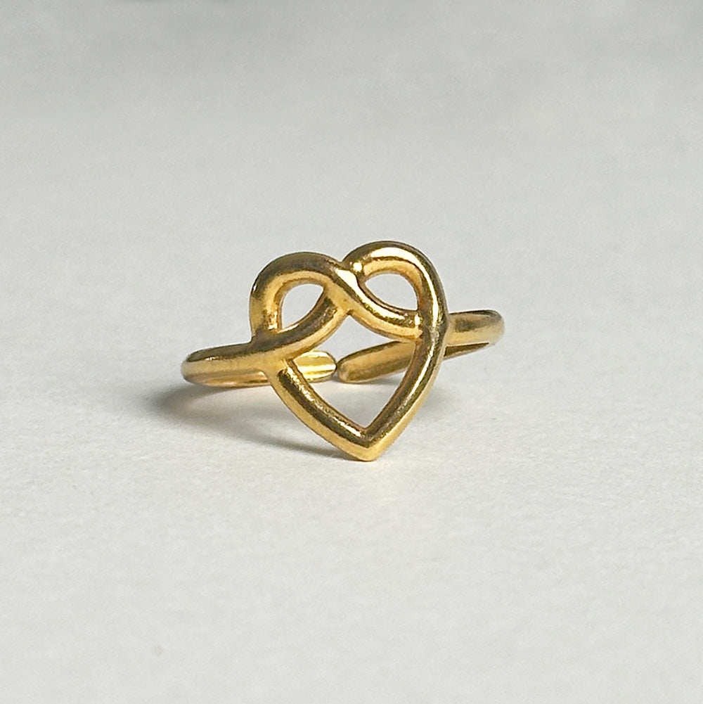 A gold heart ring featuring an interlocking heart and infinity symbol design, photographed on a neutral light gray background.