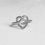 A silver ring featuring an open heart design that intertwines with an infinity symbol, photographed on a neutral light gray background.