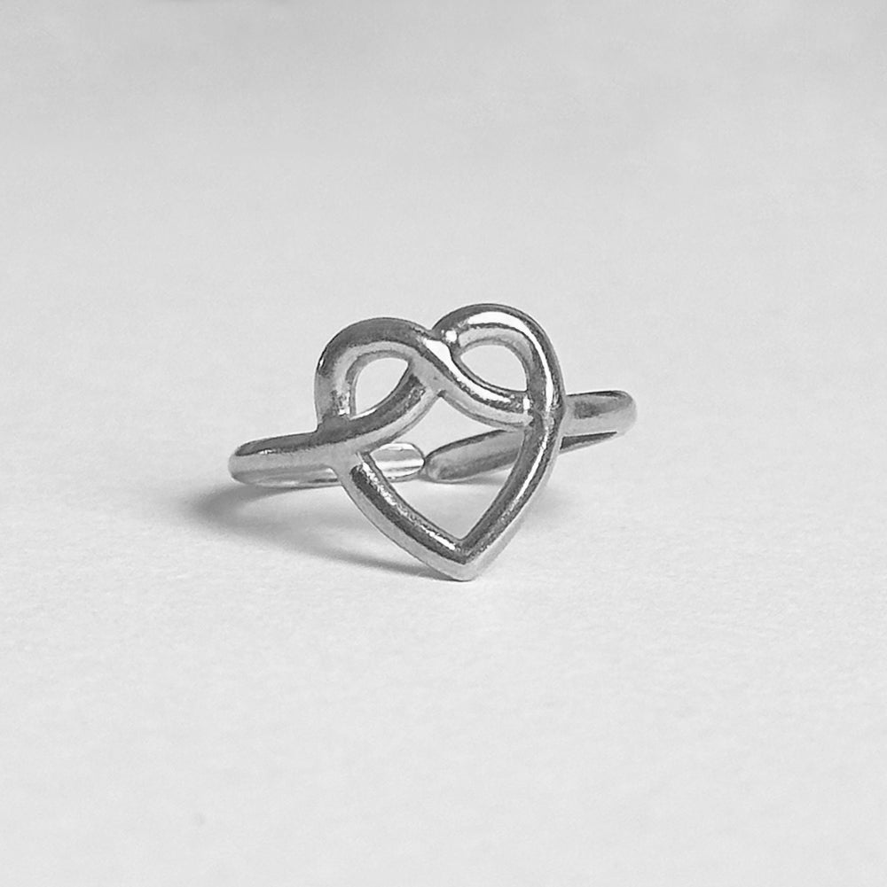 A silver ring featuring an open heart design that intertwines with an infinity symbol, photographed on a neutral light gray background.