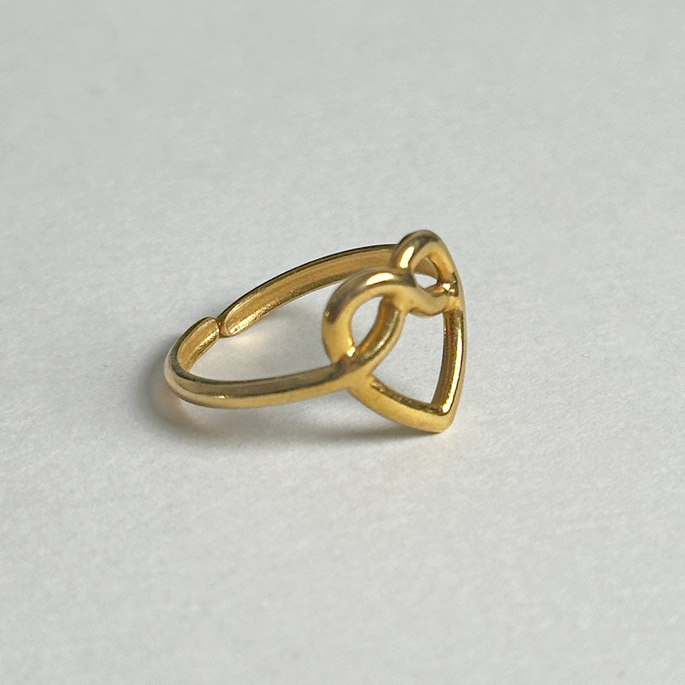 A gold ring with a heart openwork design and a central loop, photographed on a light gray textured background.