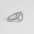 A silver open heart ring with a triangular heart shape and a small central knot detail, shown on a light gray textured background.