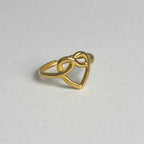 A gold ring featuring an open heart design that intertwines with an infinity symbol, photographed on a neutral light gray textured background.