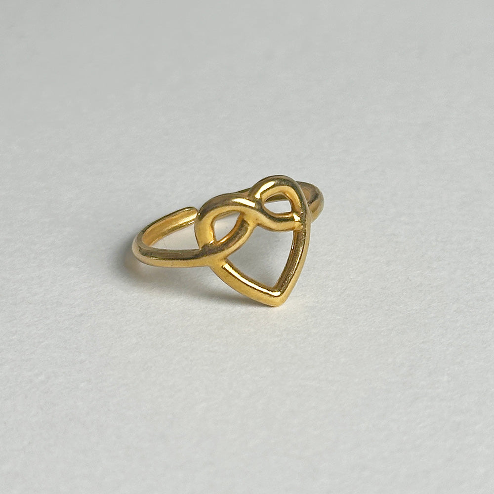 A gold ring featuring an open heart design that intertwines with an infinity symbol, photographed on a neutral light gray textured background.