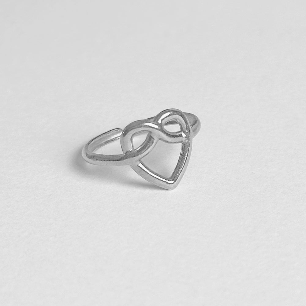 A silver ring featuring a heart-shaped openwork design with an infinity symbol at the top, resting on a light gray textured surface.