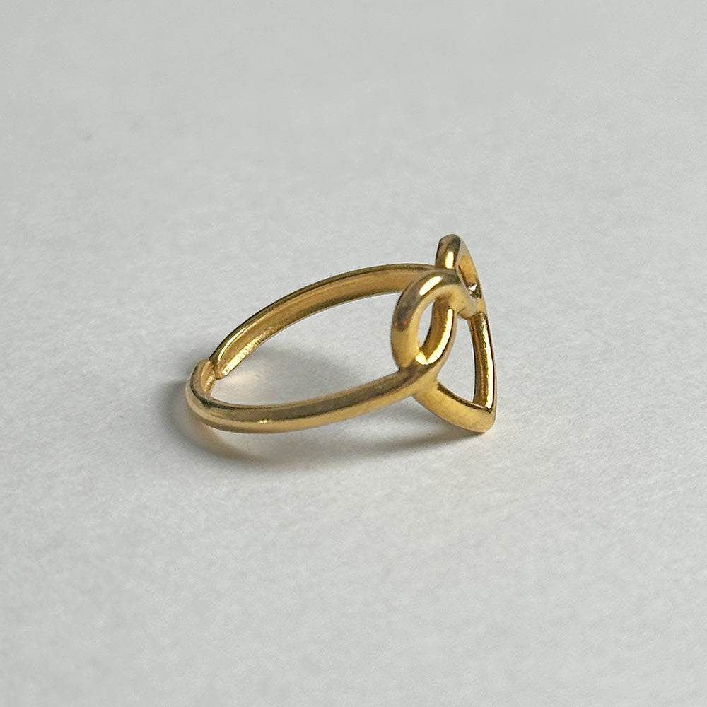 A gold ring with a minimalist triangular heart knot design, photographed on a neutral light gray background.