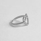 A minimalist silver ring featuring a triangular knot design, photographed on a clean, light gray textured surface.