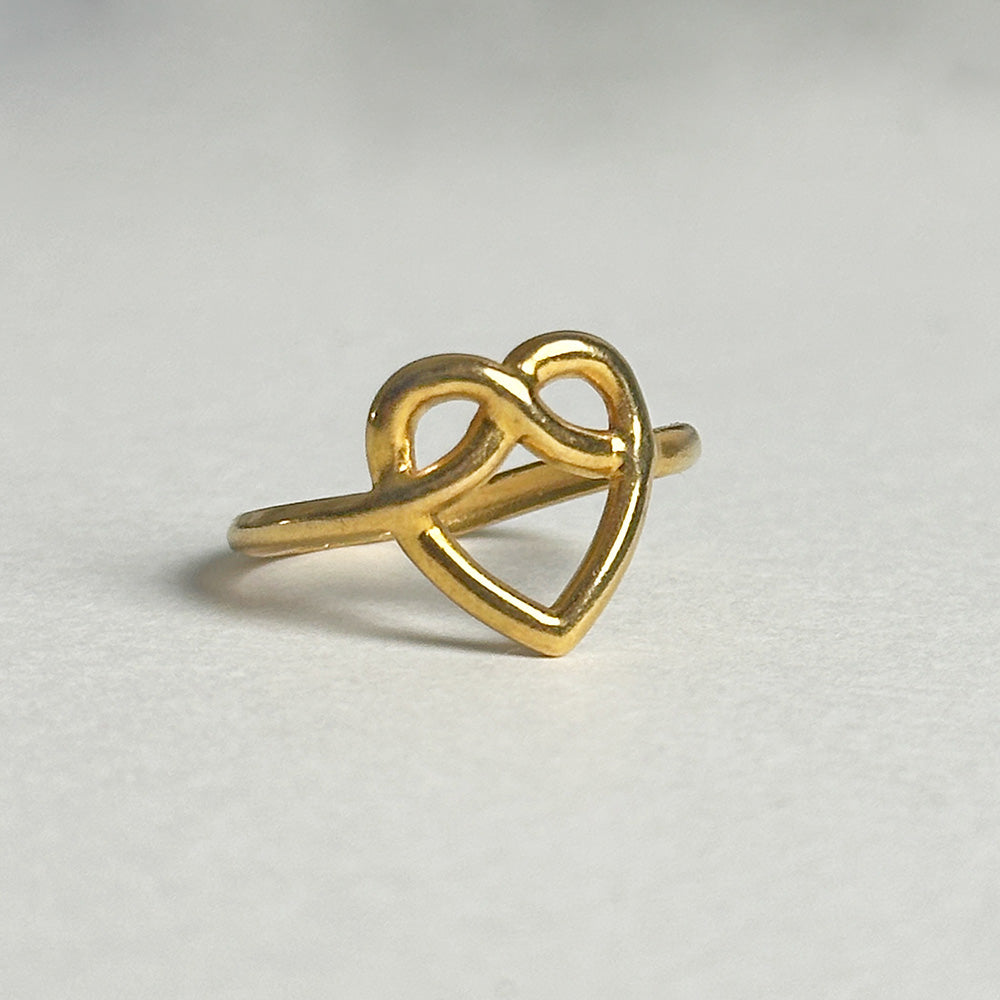 A gold ring featuring an infinity symbol that forms the outline of a heart, resting on a light gray textured surface.