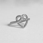 A silver ring featuring an open heart design that intertwines with an infinity symbol, photographed on a neutral light gray background.