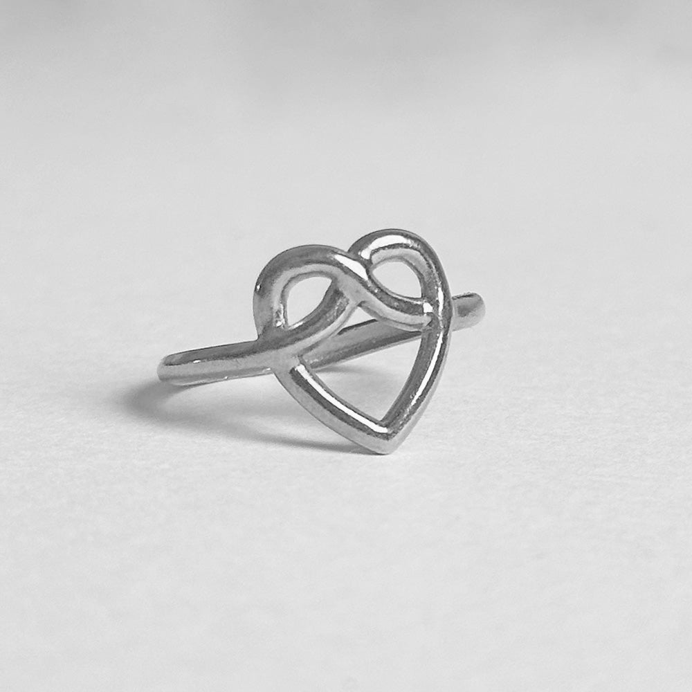 A silver ring featuring an open heart design that intertwines with an infinity symbol, photographed on a neutral light gray background.