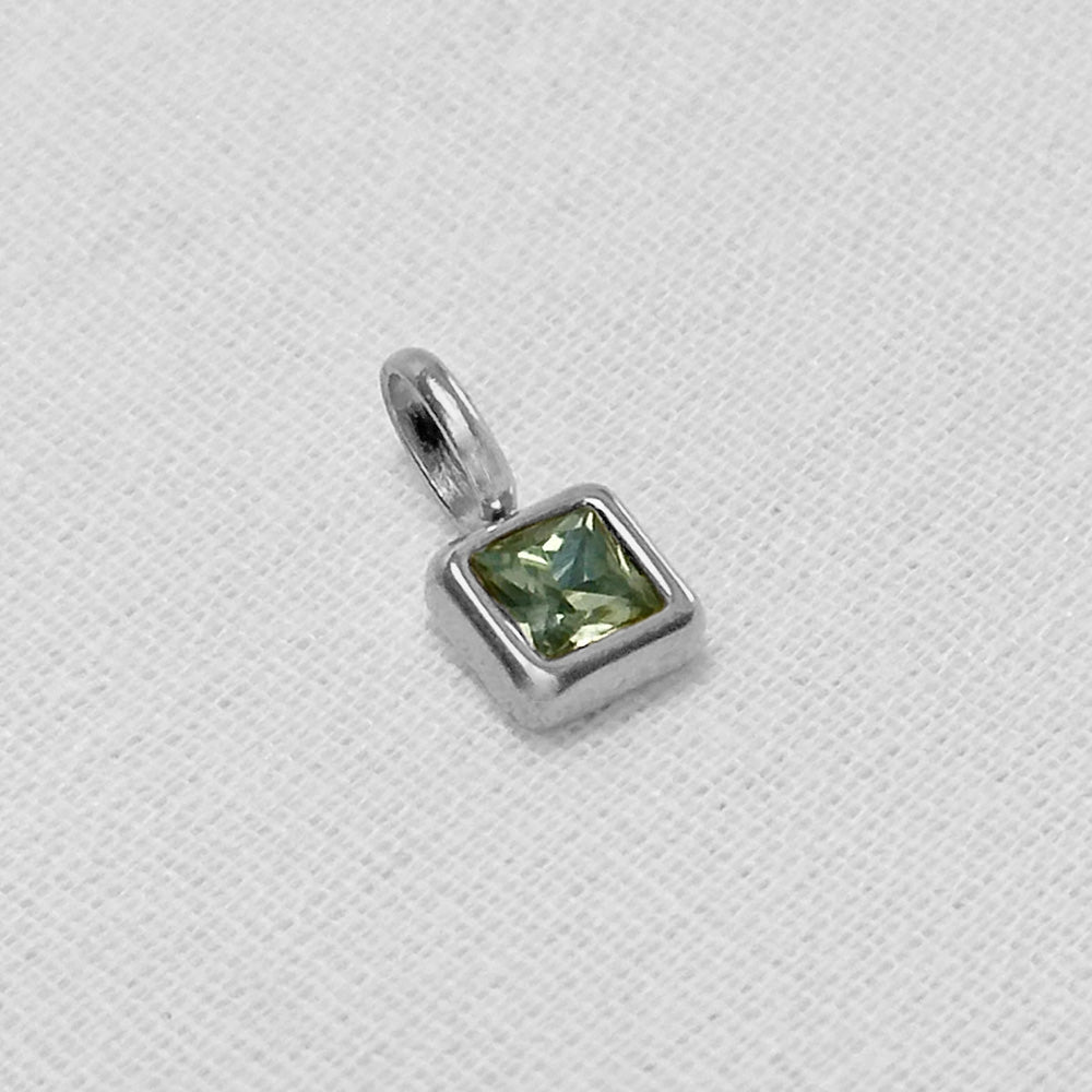 A square-cut green gemstone charm in a silver bezel setting with a small loop at the top, resting on a textured white fabric background.