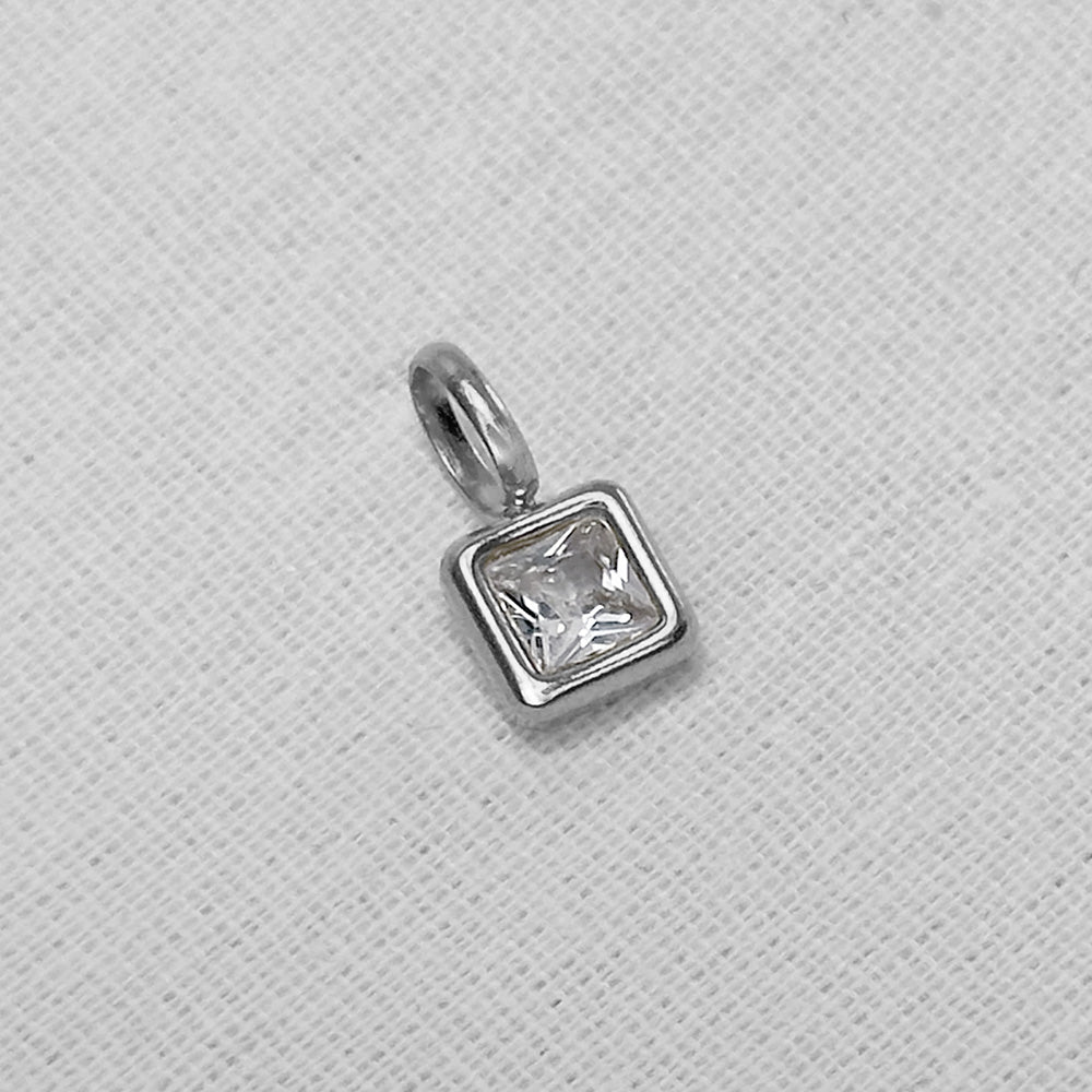A silver square charm pendant with a clear, faceted princess-cut gemstone, displayed on a textured white fabric background.