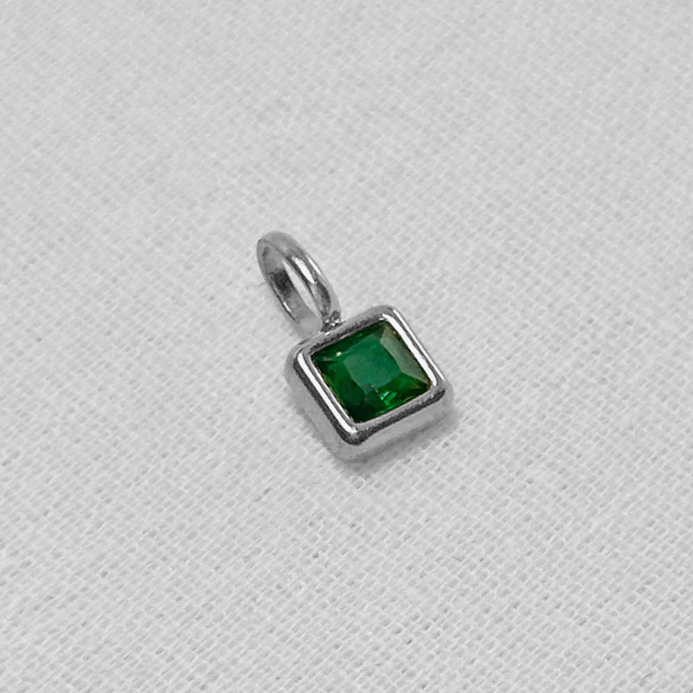 A small square pendant charm featuring a faceted green gemstone in a polished silver bezel setting with a top loop, resting on a textured white fabric background.