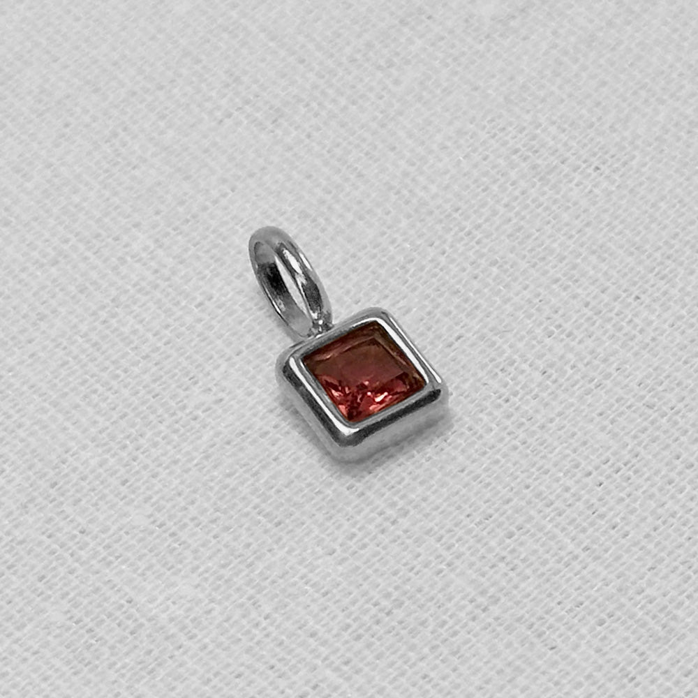 A silver square charm pendant featuring a faceted reddish-brown gemstone, displayed on a textured white fabric background.