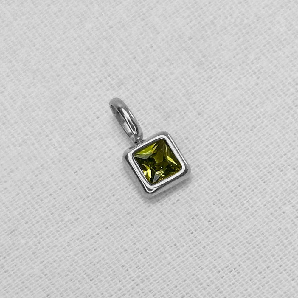 A square-cut green gemstone charm in a silver bezel setting with a silver bail, resting on a textured white fabric background.