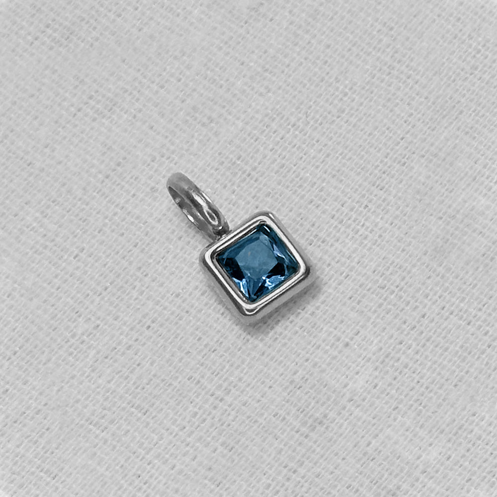 A silver charm pendant with a square-cut light blue gemstone in a bezel setting, resting on a textured white fabric background.
