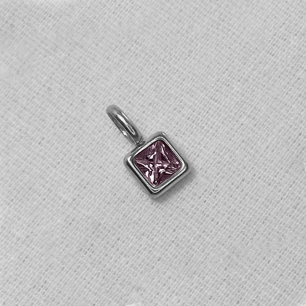 A small silver charm with a square-cut pink gemstone in a prong setting, resting on a textured white fabric background.