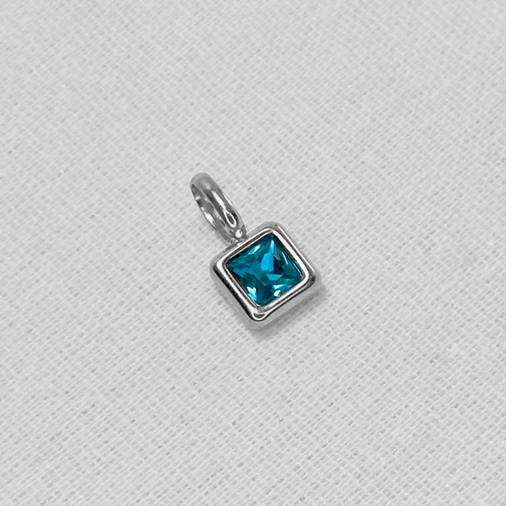 A silver square charm pendant featuring a faceted teal blue gemstone, displayed on a textured white fabric background.
