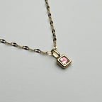 A gold chain necklace with a small square pendant featuring a pink gemstone, displayed on a light gray background.