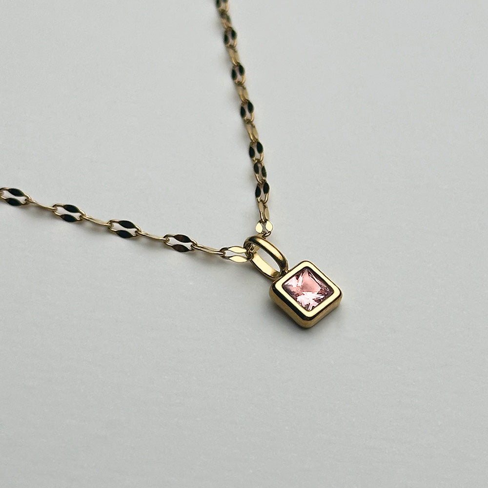 A gold chain necklace with a small square pendant featuring a pink gemstone, displayed on a light gray background.