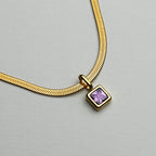 A gold snake chain necklace with a square bezel-set purple gemstone pendant, photographed on a neutral light gray background.