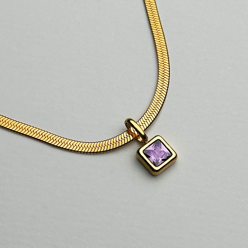 A gold snake chain necklace with a square bezel-set purple gemstone pendant, photographed on a neutral light gray background.