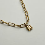 A gold paperclip chain necklace with a small square bezel-set clear crystal pendant, shown on a light gray background.