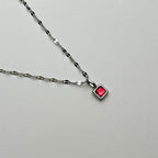 A silver chain necklace with a small square pendant featuring a vibrant red gemstone, displayed on a neutral light gray background.