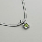 A silver snake chain necklace featuring a small square pendant with a green gemstone, displayed on a neutral light gray background.
