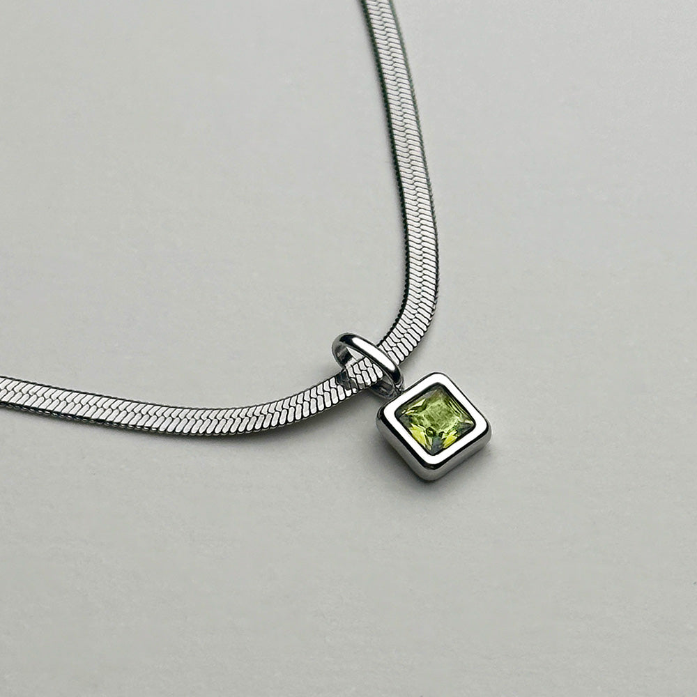 A silver snake chain necklace featuring a small square pendant with a green gemstone, displayed on a neutral light gray background.