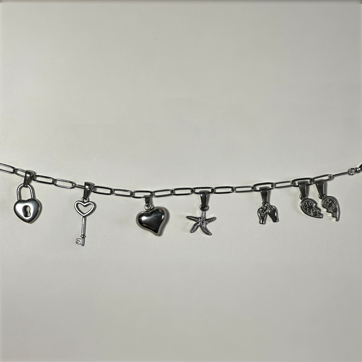 A silver paperclip chain bracelet featuring multiple dangling charms, including a heart-shaped padlock, a key, a heart, a starfish, and a couple of small animal figures, displayed against a plain light gray background.