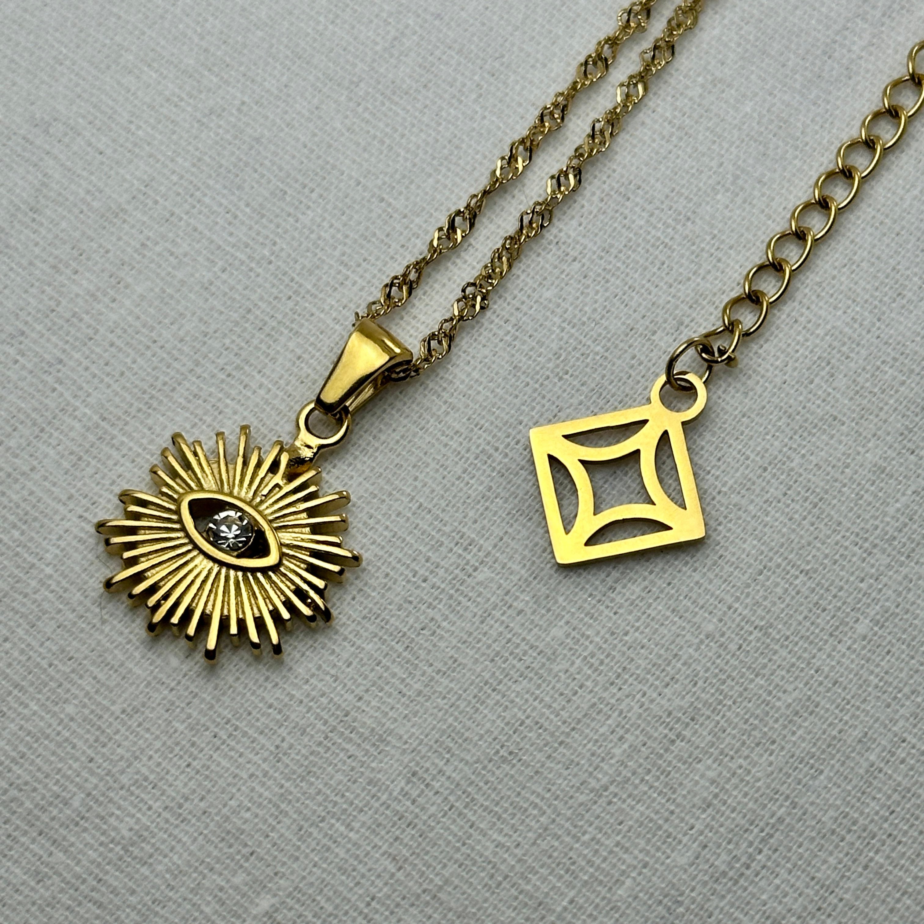 Gold necklace with a sunburst pendant featuring a central clear stone, alongside a square geometric charm.
