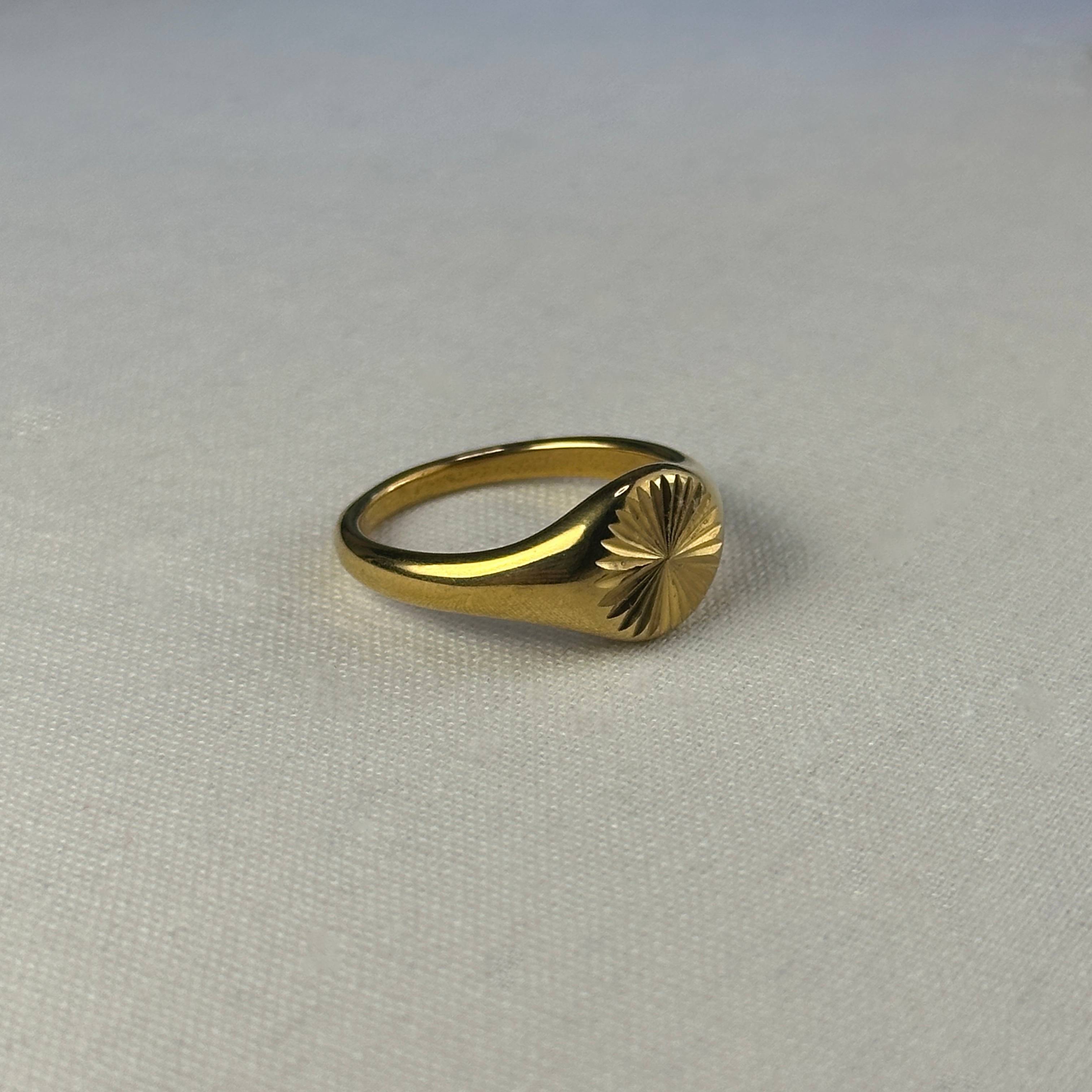 Gold signet ring with sunburst design on a textured white background.
