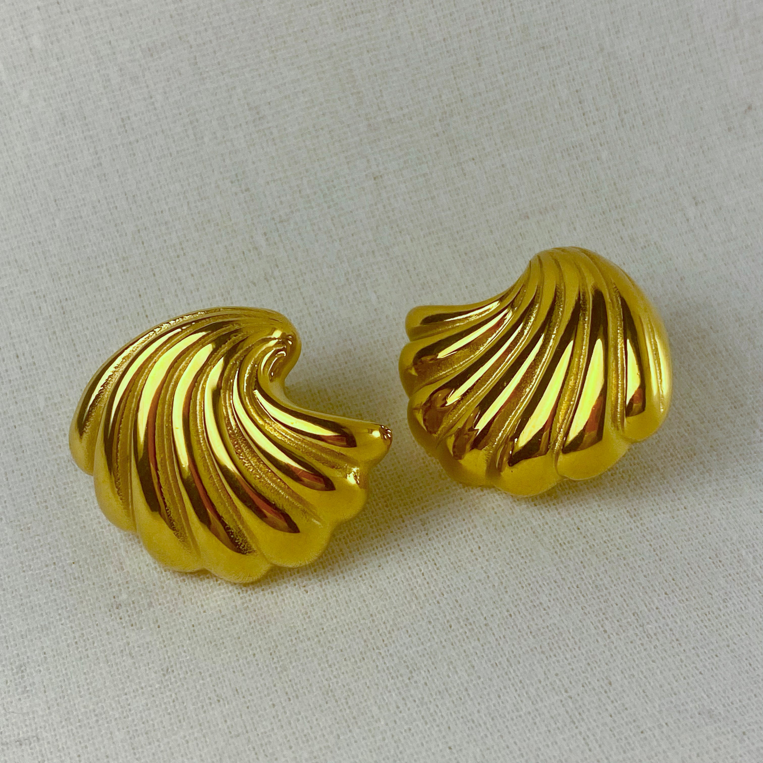Two gold, shell-shaped earrings on a white fabric background.