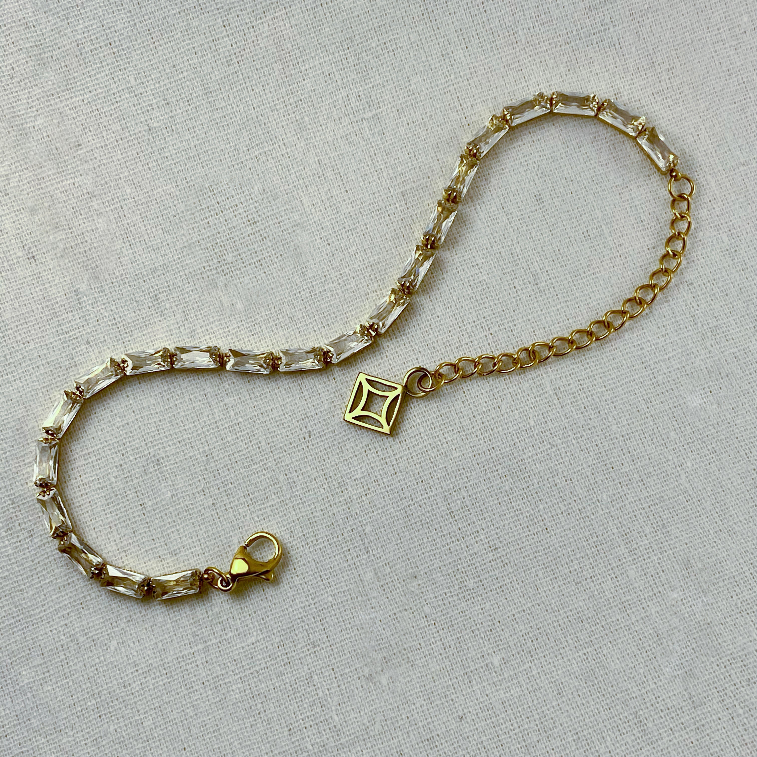 Gold bracelet with clear baguette crystals and a square charm on a light-colored linen background.
