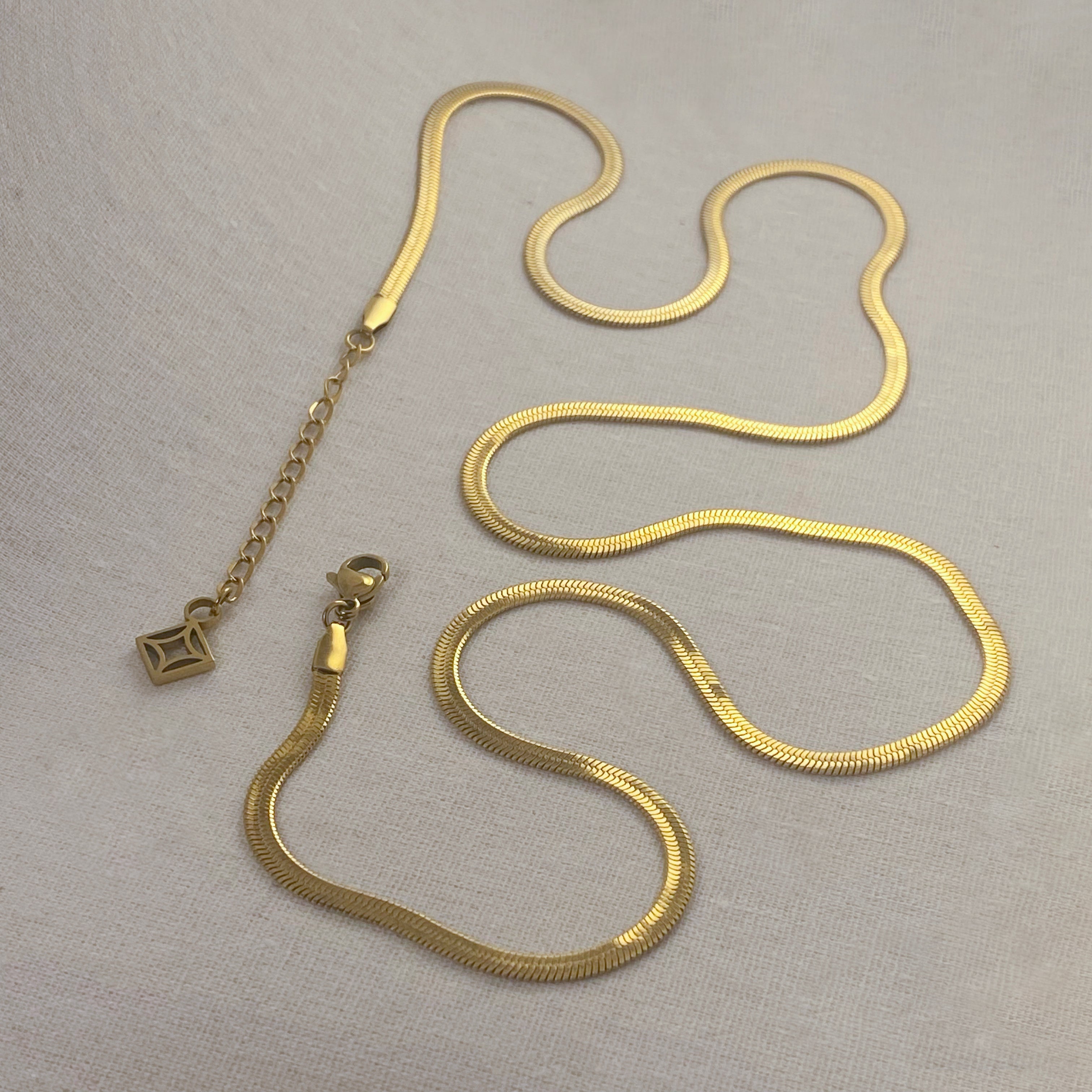 Close-up of gold-toned, herringbone chain-link necklace and bracelet on a textured cream background.