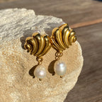 Gold shell-shaped earrings with dangling white pearls on beige stone background
