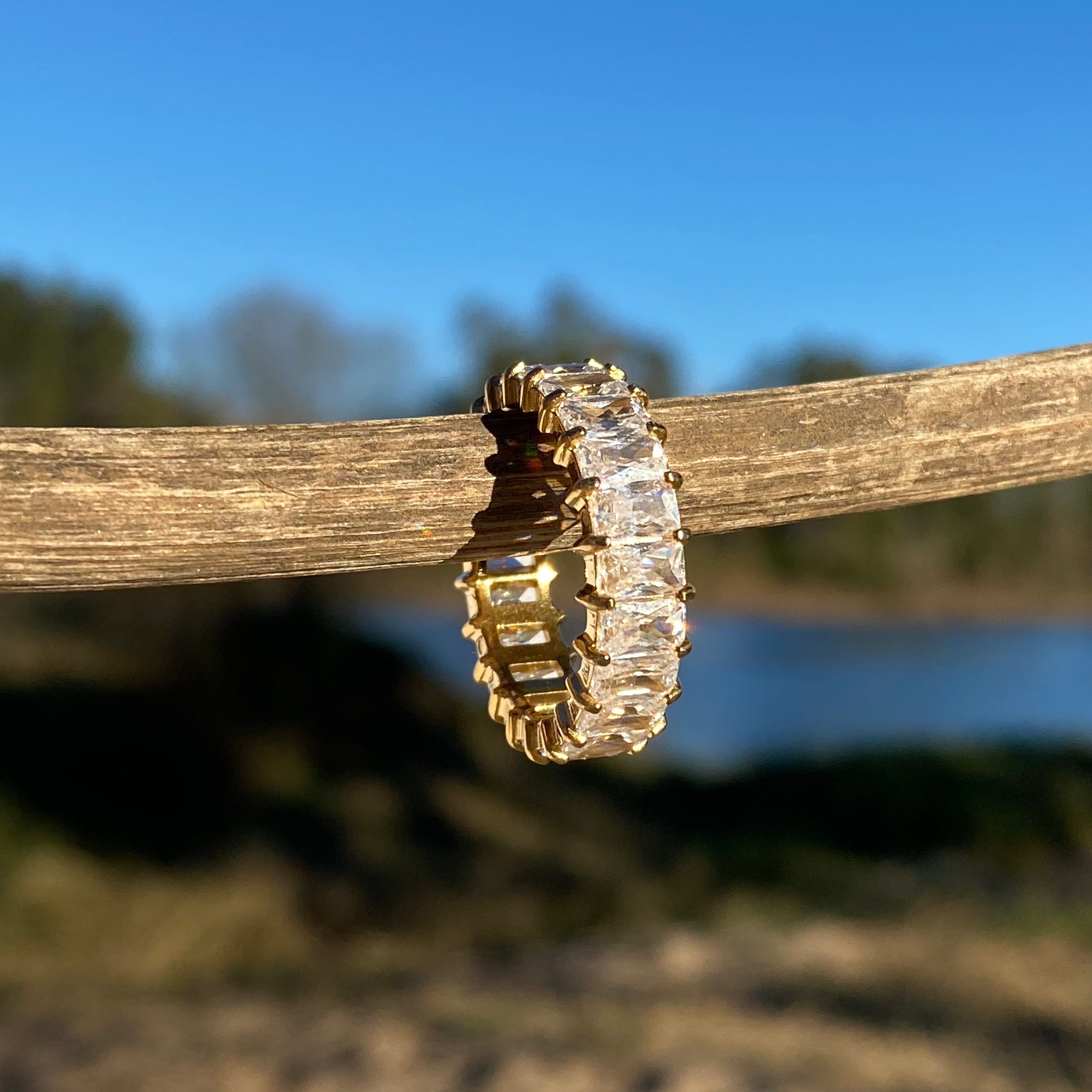Gold eternity ring with emerald-cut clear stones on a wooden branch with a blue sky and blurred nature background.