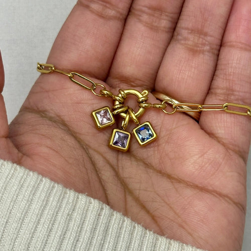 Hand holding gold bracelet with three square gemstone charms in pink and blue