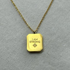Gold pendant necklace with engraved text 'I AM STRONG' on textured light fabric background