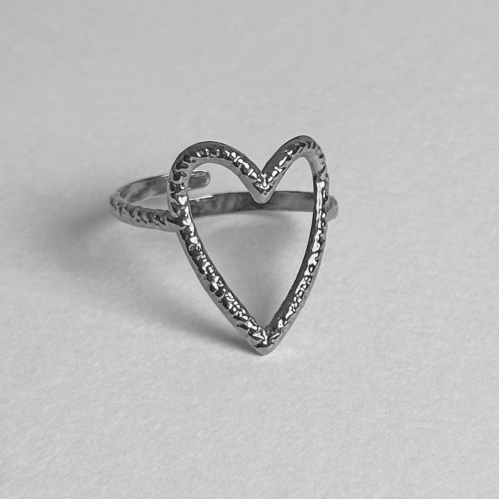 A silver ring featuring an open heart design with a textured, hammered finish on the heart outline and a smooth, beaded band, shown on a light gray background.